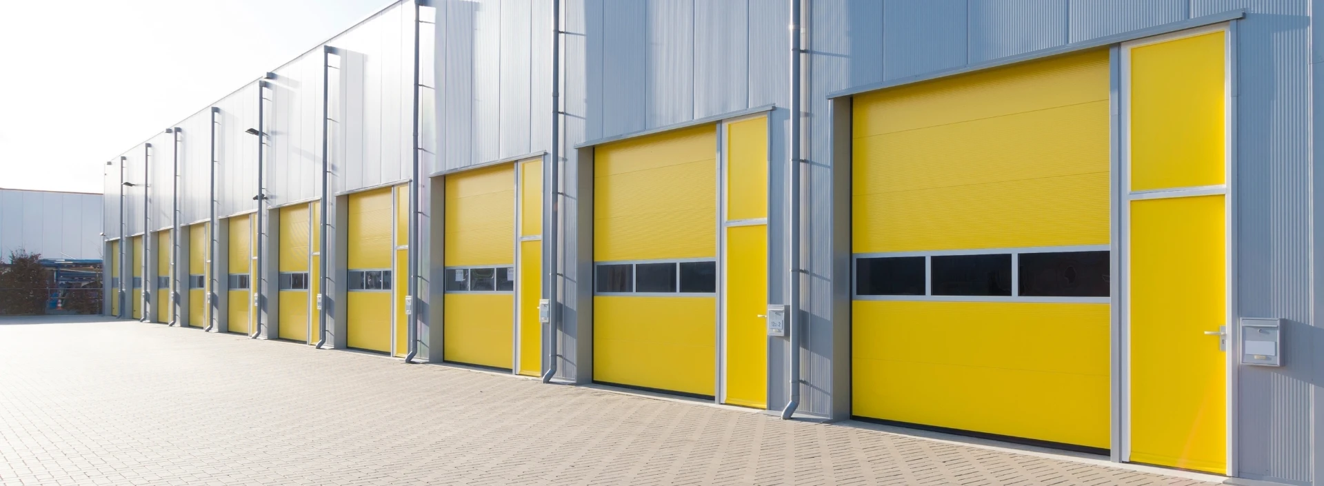 A row of seven identical industrial garage doors in yellow, featuring metal frames, at a commercial building stands out. They are fitted with small rectangular windows in the middle. The ground is paved, and nearby signs hint at guidelines akin to those found in a detailed Theline Painting FAQ.