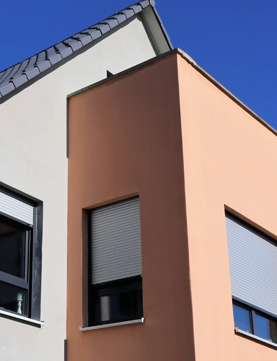 Modern two-story building facade with beige and white walls, two windows with closed shutters, and a tiled roof under a clear blue sky, perfectly showcasing the quality of New Construction Painting Services.